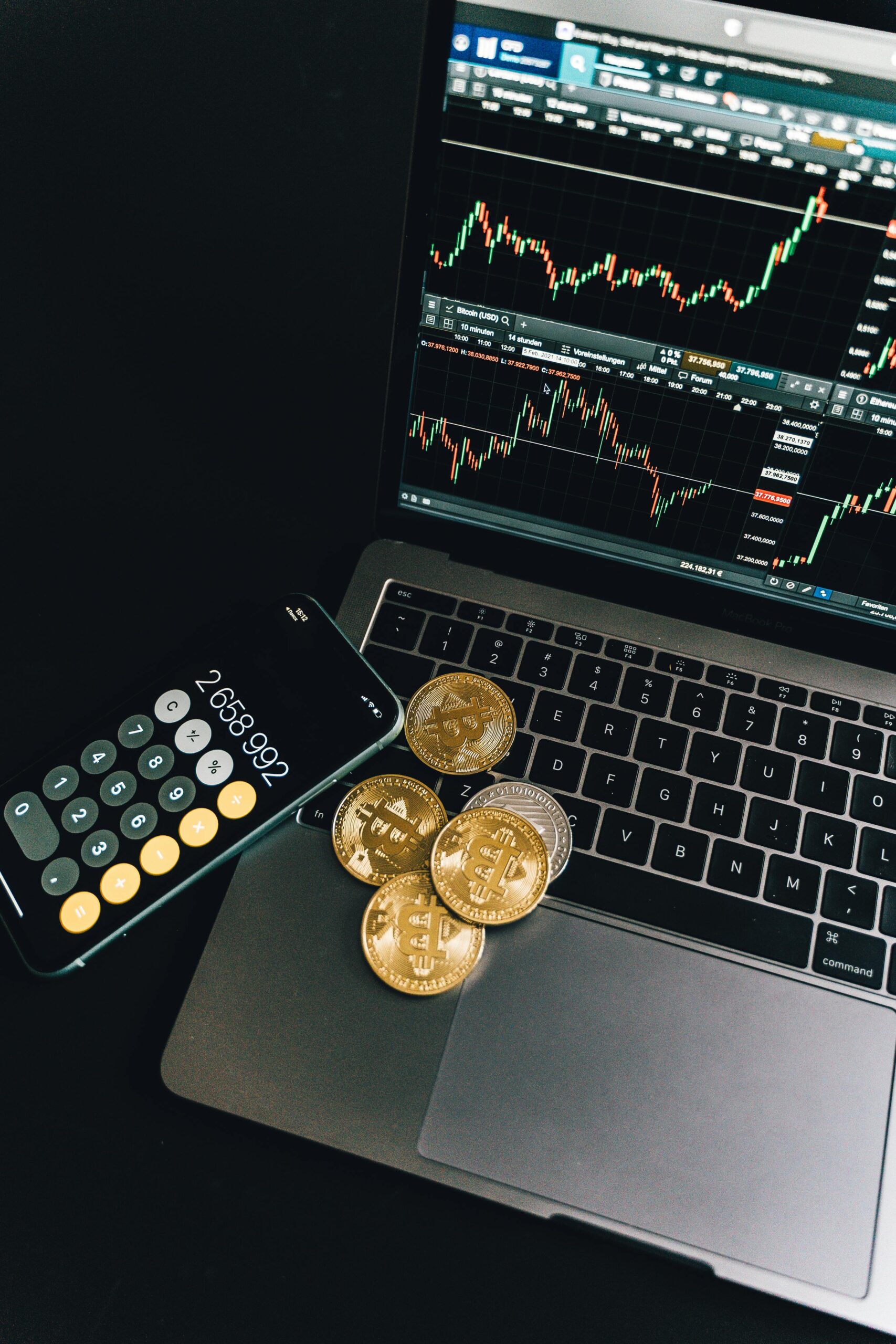 A still life of Bitcoin coins, a smartphone calculator, and a trading chart on a laptop.